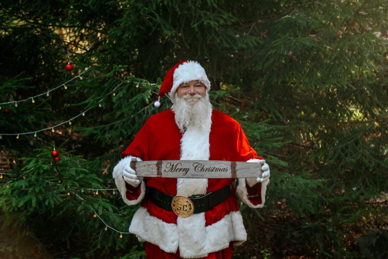 Santa Brian holding a Merry Christmas sign among evergreen trees